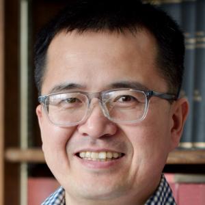 Junpeng Wang wearing a blue plaid shirt and glasses smiling for his headshot in front of a bookshelf.