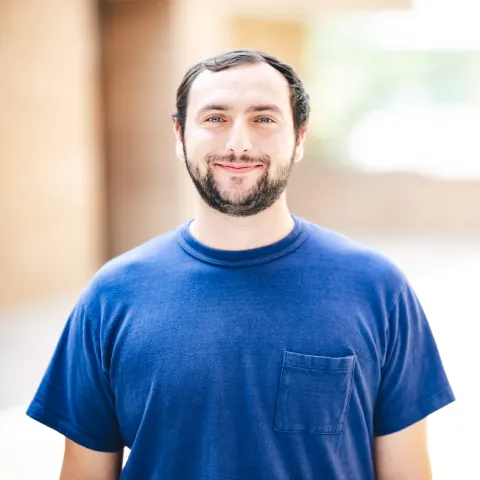Avery Stamps wearing a blue t-shirt smiling in front of a brick building for his headshot.