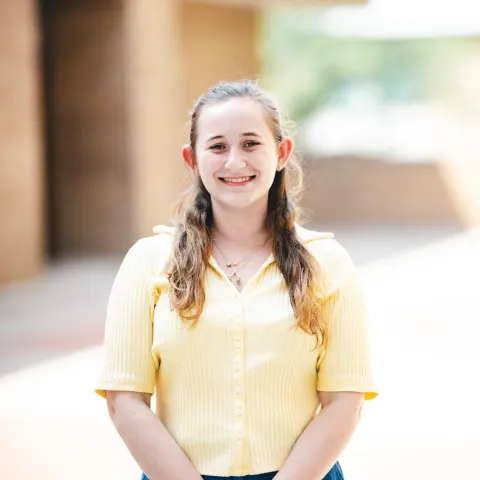 Daphne Schencavitz wearing a yellow shirt smiling in front of a brick building for her headshot.