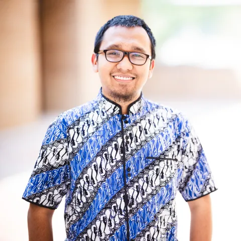 Herbi Yuliantoro wearing a blue shirt with designs smiling for his headshot in front of a brick building.