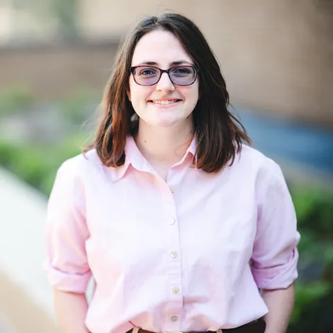 Isabelle Herlinger standing outside in front of a brick building wearing a pink shirt for her headshot.