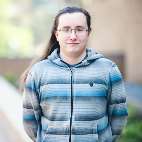 Jordan Polvere wearing a striped hooded zip up jacket smiling outside for his headshot.