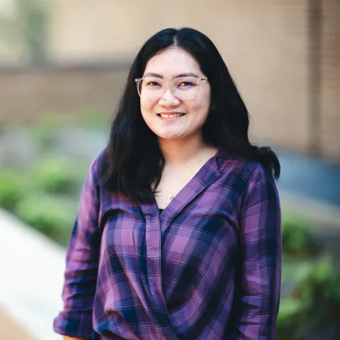Korina Vida Sinad standing in front of a brick building wearing a purple and black button-up shirt for her headshot.