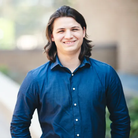Luc Mauro standing in front of a brick building wearing a blue button-up shirt for his headshot.