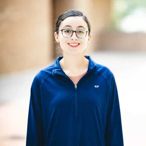 Lyndsey Buren wearing a blue zip up jacket and glasses smiling for her headshot in front of a brick building.