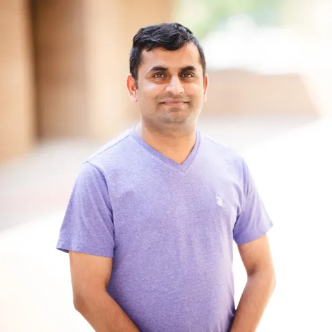 M Iftikhar wearing a purple shirt smiling for his headshot in front of a brick building