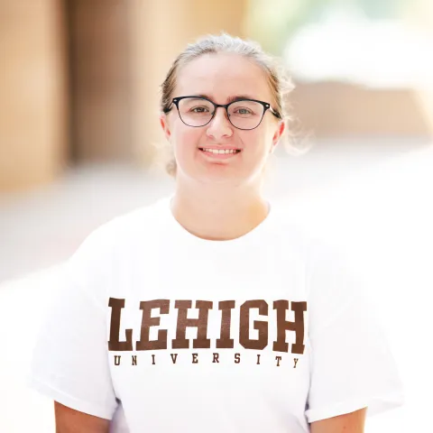 Madison Pellerito wearing a white Lehigh t-shirt and glasses smiling for her headshot in front of a brick building.