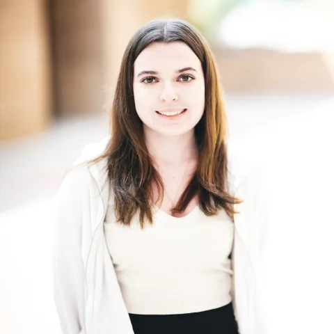 Patricia Olsen wearing a white top and white zip up smiling for her headshot in front of a brick building.