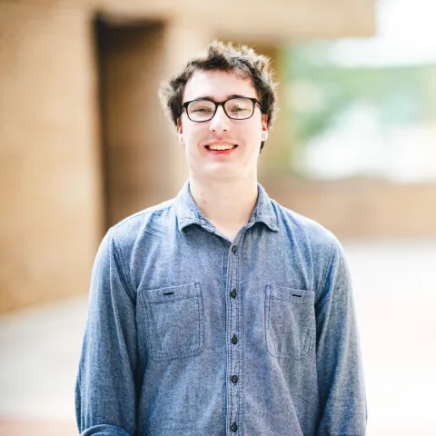 Wesley Rauenzahn wearing a blue shirt and glasses smiling for his headshot in front of a brick building.