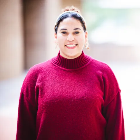 Yudeliz Sanchez wearing a pink sweater smiling for her headshot in front of a brick building.