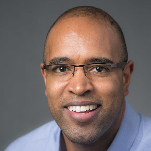 Professor Jebrell Glover wearing a blue button down shirt in front of a gray background