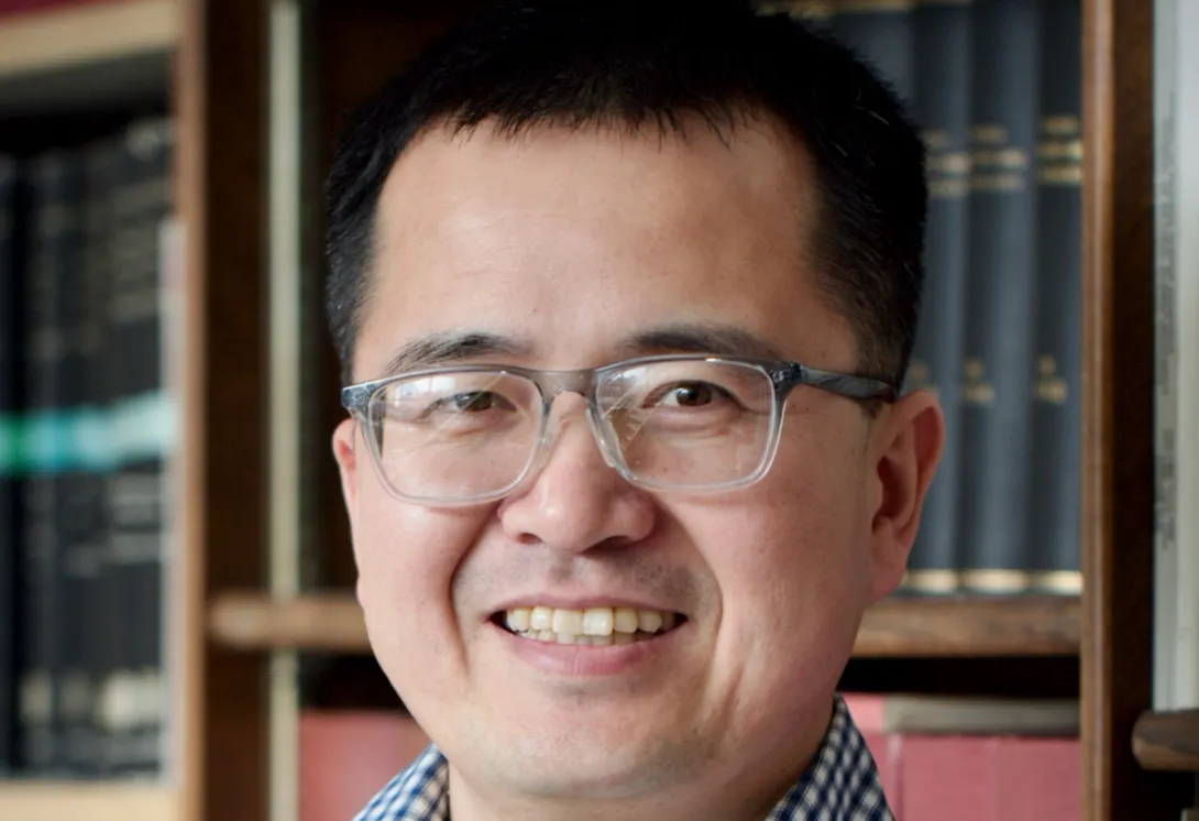 Junpeng Wang wearing a blue plaid shirt and glasses smiling for his headshot in front of a bookshelf.